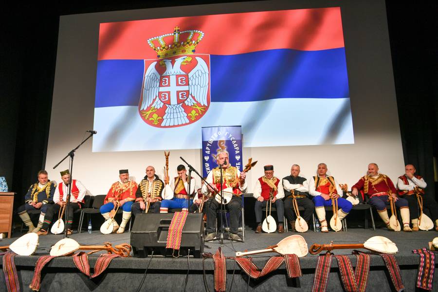 Гуслари на сцени. Један гусла у првом плану, остали седе иза њега и слушају га. Сцена је украшена појасевима и гуслама. На позадини је застава Републике Србије (црвена, плава и бела са грбом двоглавог орла са круном)