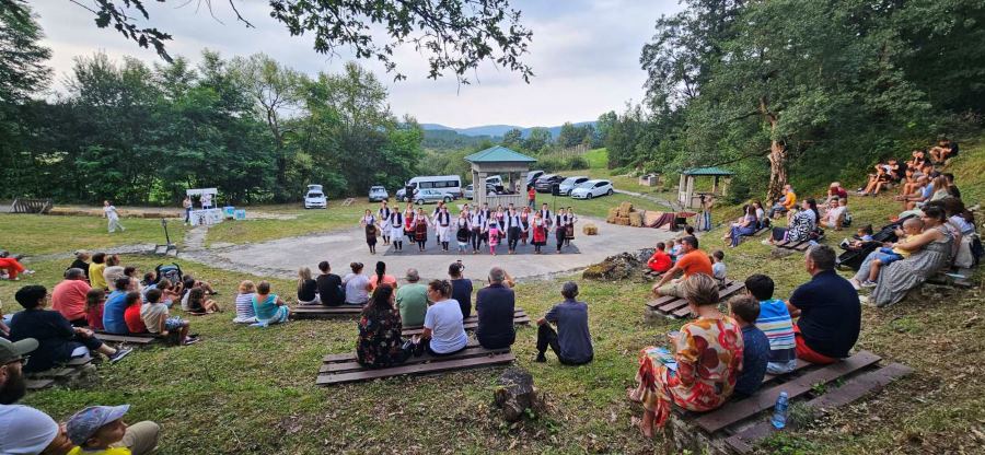 Letnja pozornica u selu tripkova. Ljudi sede na klupama u prirodi i gledaju nastup Kulturno-umetnickog drustva