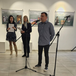 Човек говори испред микрофона. У позадини две жене