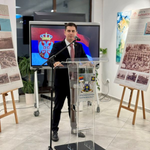 Covek govori za govornicom iza njega je televizor sa zastavom