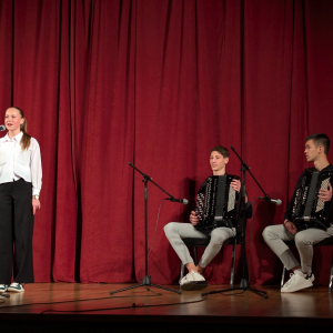 devojcica stoji i peva, iza nje sede decaci sa harmonikama