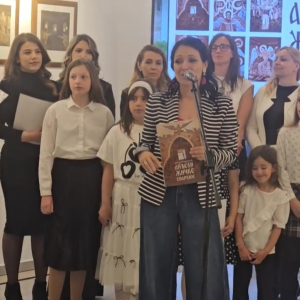 zena stoji i prica, iza nje deca i odrasli 