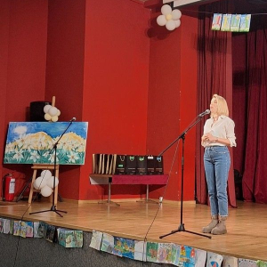 zena stoji beloj kosulji i farmerkama i prica na mikrofon