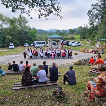 Letnja pozornica u selu tripkova. Ljudi sede na klupama u prirodi i gledaju nastup Kulturno-umetnickog drustva